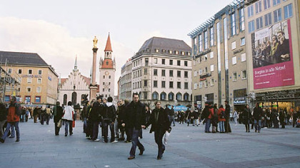 Bustling Marienplatz with its municipal buildings, offices and stores, is the center of Munich. A gilded statue of the Virgin Mary stands in the middle of the plaza. At one end is the old city hall, with its distinctive red roof.